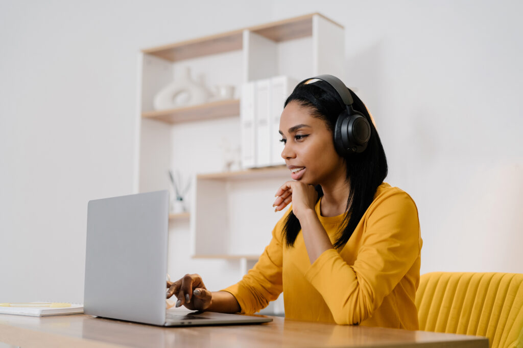 A woman wearing headphones sits at her laptop in a bright, modern workspace, appearing confident and focused as she works on her podcast setup and prepares to speak with authority. The clean desk and recording gear highlight the professional, polished side of building a podcast that strengthens her brand presence.