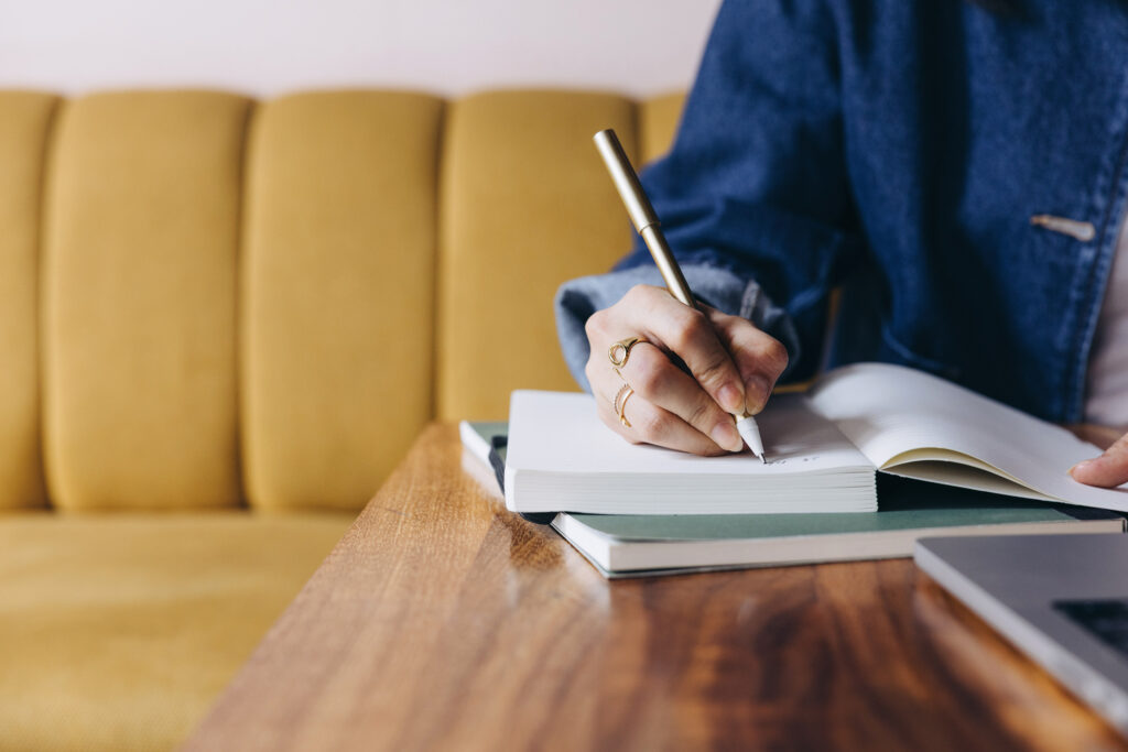 A person writing notes in a journal at a desk with a laptop nearby, symbolizing planning and refining a podcast strategy when a show isn’t growing and needs clearer direction.