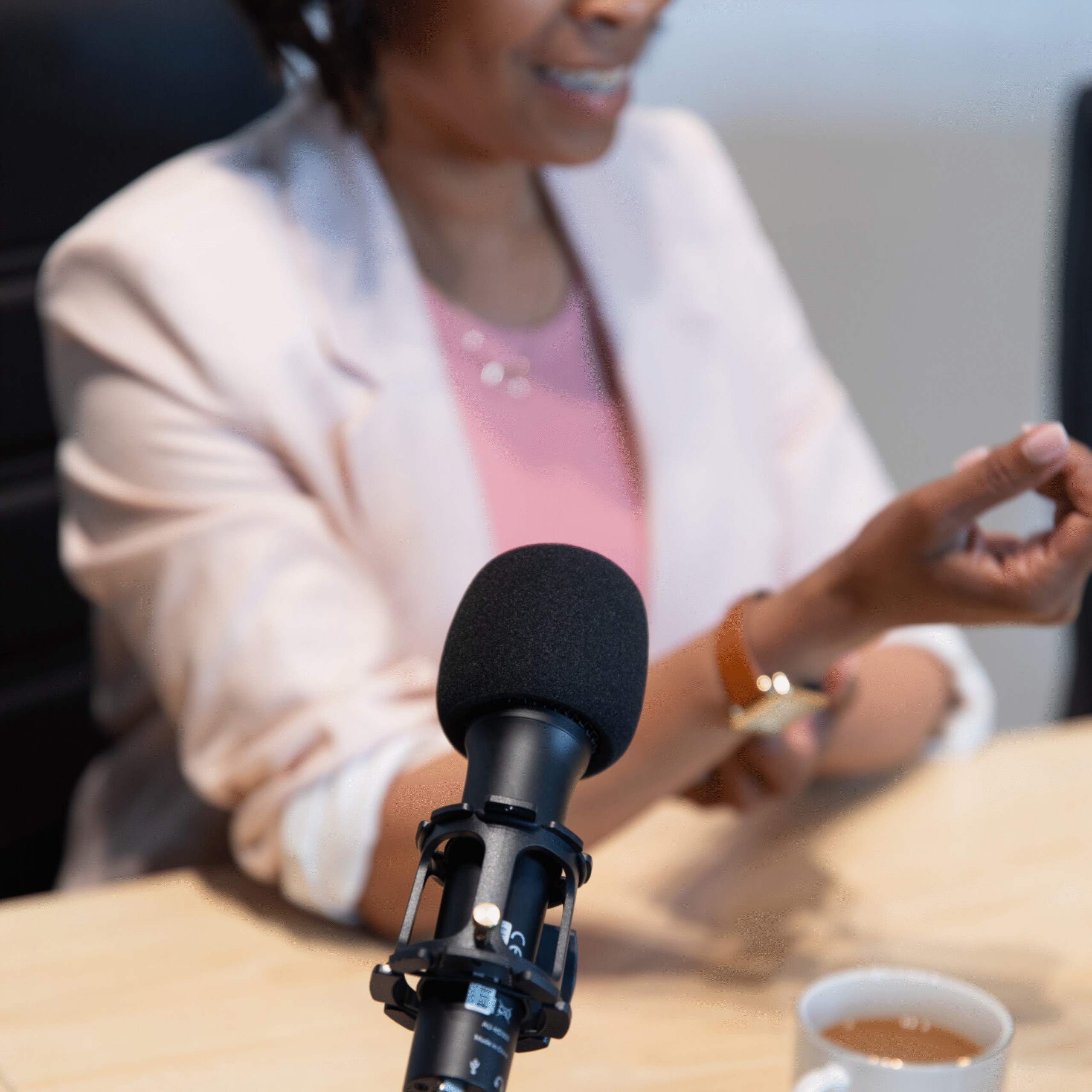 podcast-visibillity-strategies Close-up of a podcast microphone on a desk in front of a woman in a pink blazer, speaking during a podcast recording.