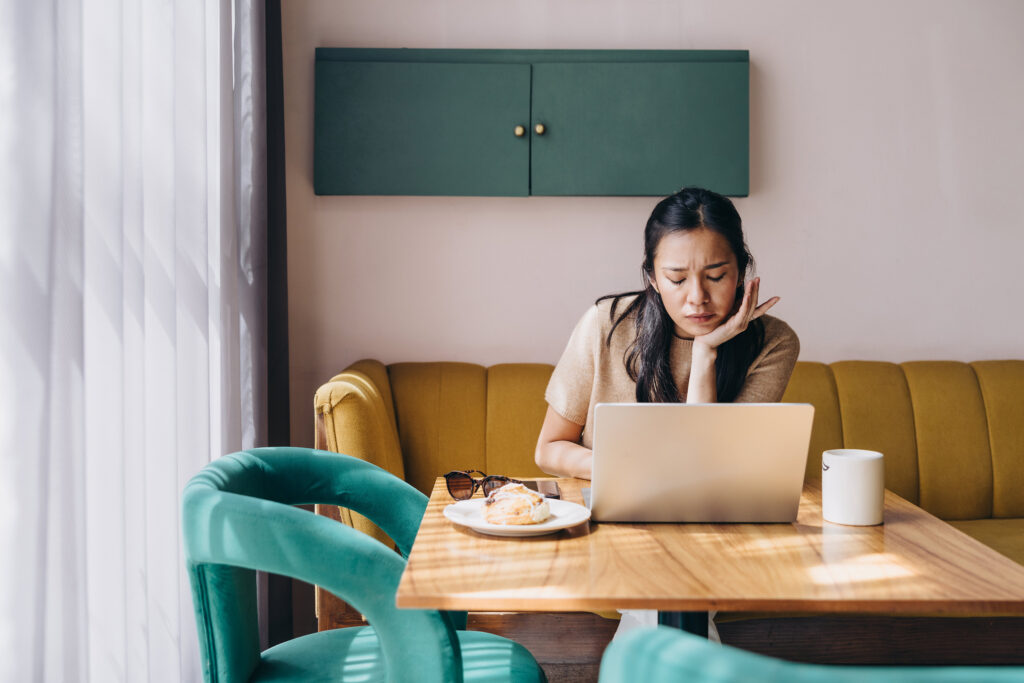 A woman sits in a sunlit café, leaning over her laptop with a frustrated, contemplative expression, as if troubleshooting her podcast strategy. A pastry, coffee mug, and sunglasses sit on the table, capturing the “working hard but not seeing results” moment many podcasters experience.