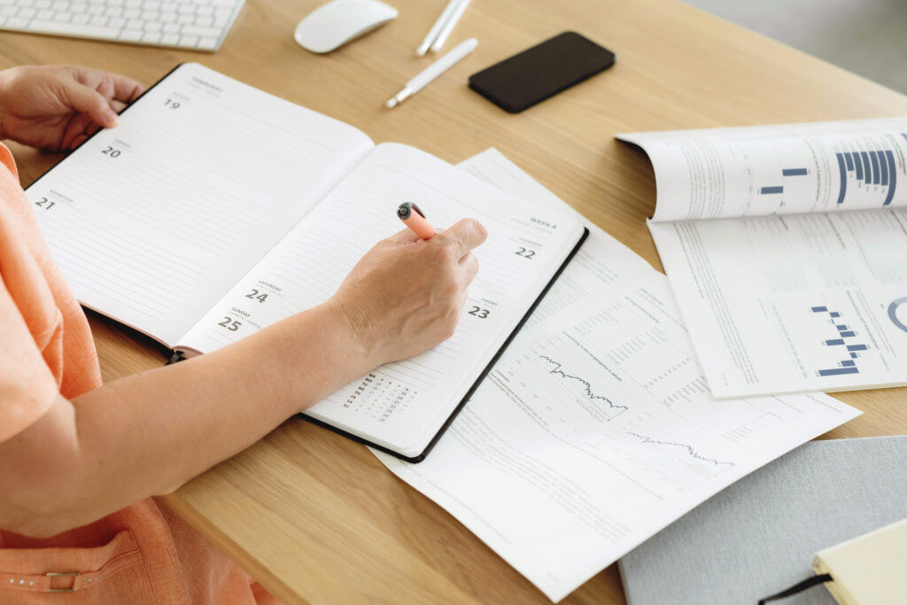 A person writes in a content calendar during a planning session at an organized workspace, surrounded by charts and documents that reflect a strategic, intentional approach to managing projects and scheduling upcoming content.