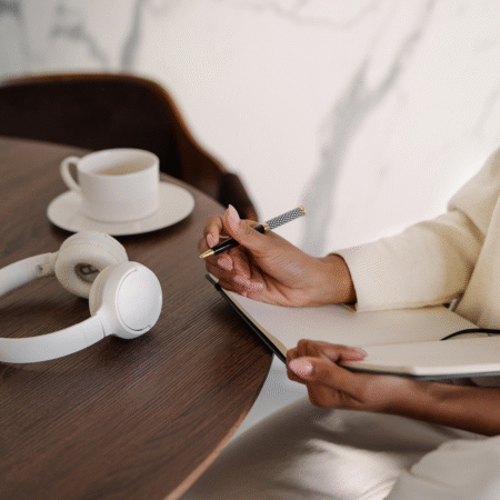 how-to-write-podcast-titles-that-get-you-found-blog Woman writing podcast episode titles in a notebook with headphones and coffee on the table — planning clear, SEO-friendly podcast content
