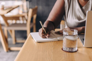 Close-up of a woman writing notes in a notebook beside a laptop and glass of water, representing podcast content planning and business strategy development.