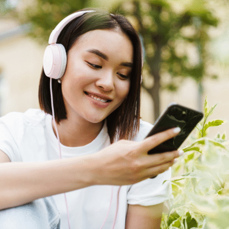 Woman listening to a podcast on her phone with pink headphones outdoors, representing podcast SEO and discoverability for entrepreneurs.