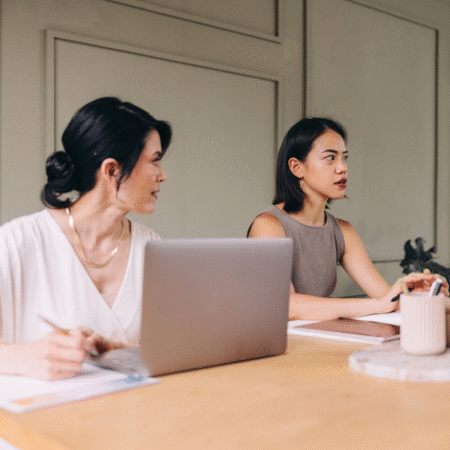 Two women in a meeting discussing strategy, one working on a laptop and the other speaking, representing the importance of a clear podcast workflow and intentional planning for better podcast growth.