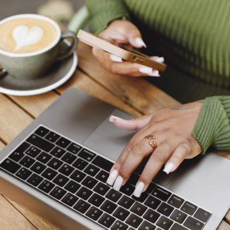 Hands typing on a laptop while holding a smartphone next to a latte, representing podcast strategy work and content planning for entrepreneurs supported by Leah Bryant Co.