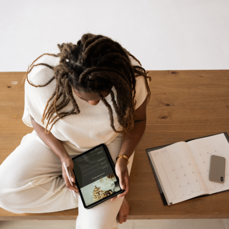 A woman sitting at a wooden table reviewing content on a tablet while a planner and phone sit beside her. She appears to be planning or organizing her podcast workflow, symbolizing productivity, content strategy, and episode planning.
