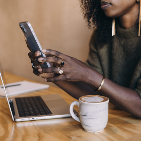 Black woman podcaster reviewing podcast strategy on phone with laptop and coffee, checking podcast listener data for business growth