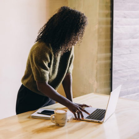 A woman working on a laptop at a wooden desk with a coffee cup and notebook, researching podcast discoverability strategies.