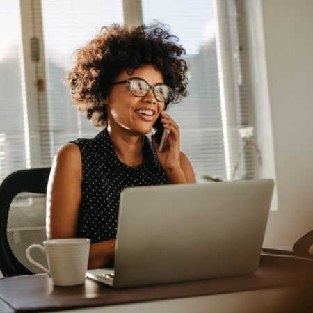 Woman with curly hair and glasses smiling while on a phone call at her laptop — podcast conversion strategy for business owners