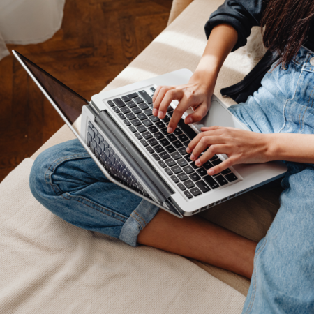 Woman sitting cross-legged on a couch working on a laptop, representing podcast consistency and strategic podcasting for business growth