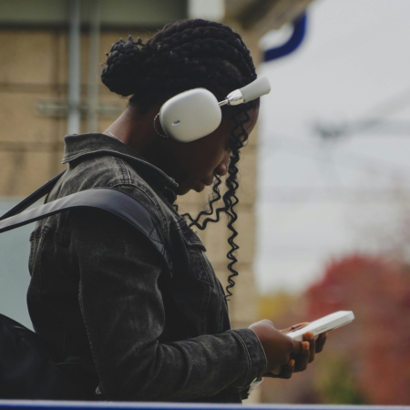 Woman wearing white over-ear headphones looking at her phone, representing podcast discoverability and how listeners search for and find new podcasts on their mobile device.
