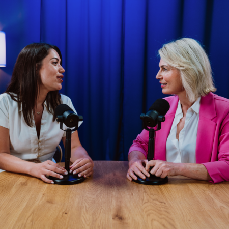 Two women sitting across from each other at a wooden table with podcast microphones, discussing podcast guesting strategy in a professional studio setting
