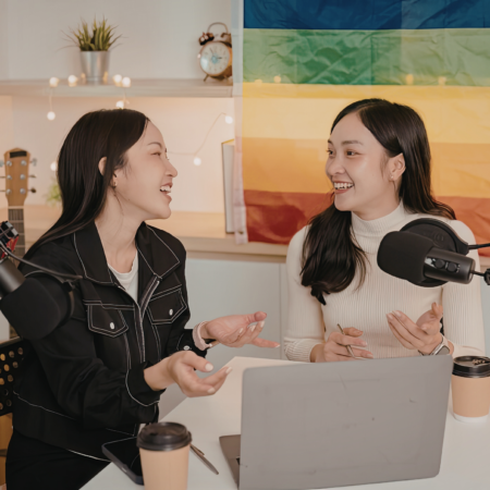 Two women smiling and talking at a podcast recording setup with microphones, a laptop, and coffee cups on the table, discussing podcast success metrics for business owners.