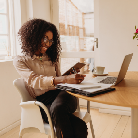 Woman sitting at a home office desk reviewing content on a tablet with a laptop and coffee nearby, representing podcast content strategy for business owners.