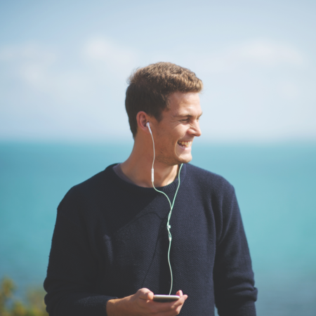 Man listening to podcast outdoors in summer with wired earbuds and smartphone near water