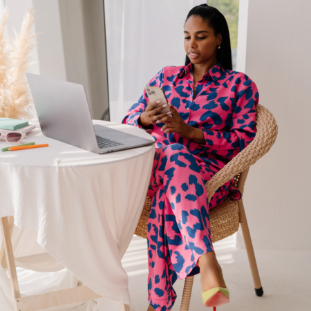 Read the full post on how to convert podcast listeners to clients. Woman in a pink and blue leopard print shirt and pants sitting in a woven chair at a small white table, checking her phone with a laptop open in front of her.
