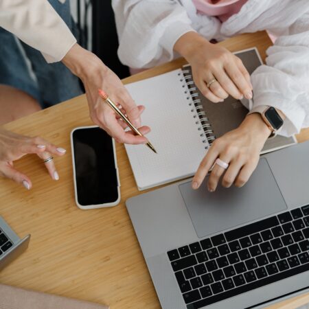 Two women collaborating at a wooden desk with a laptop, notebook, and phone, one holding a gold pen while the other points at the laptop screen during a coaching session.
