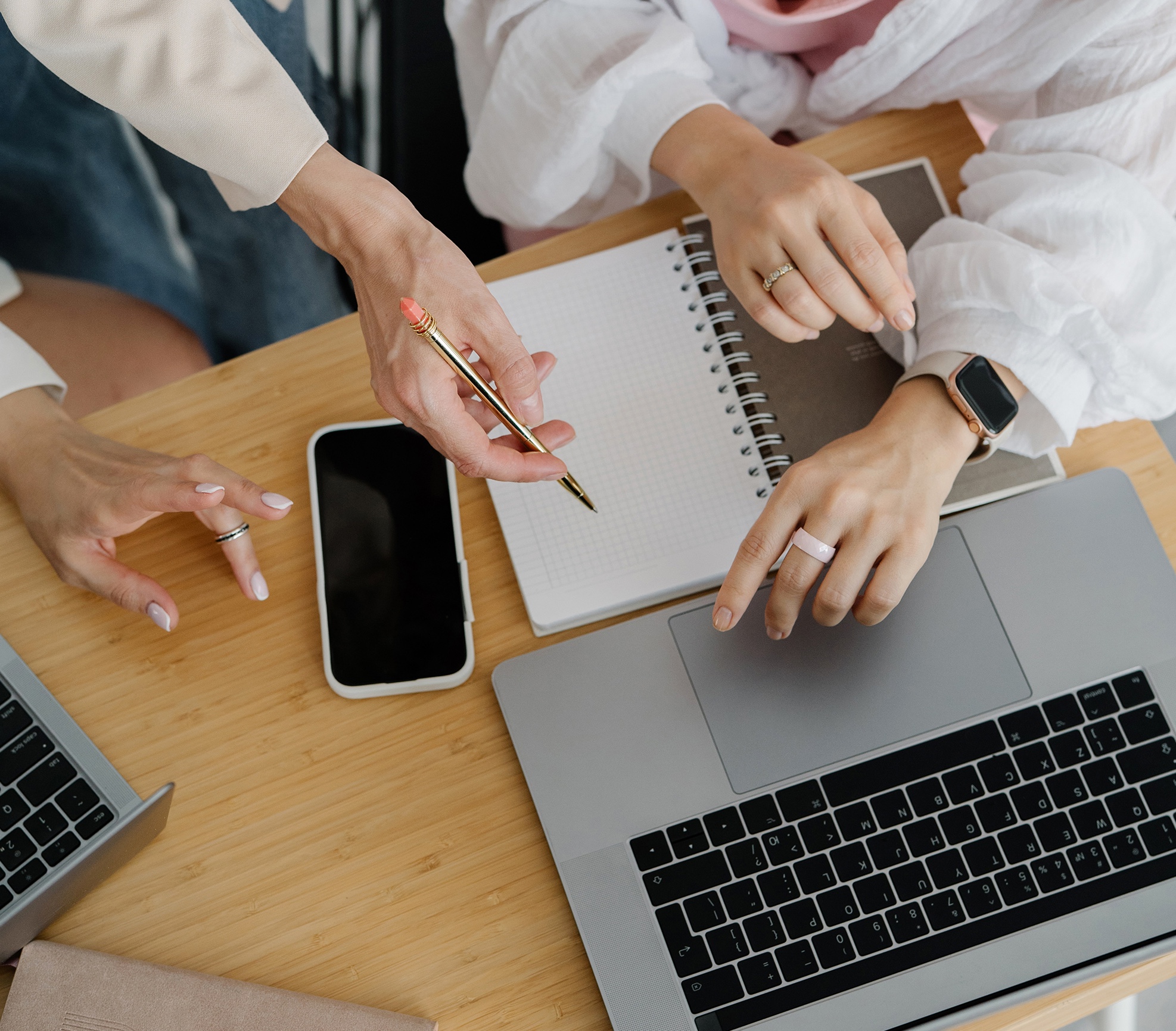 Two women collaborating at a wooden desk with a laptop, notebook, and phone, one holding a gold pen while the other points at the laptop screen during a coaching session.