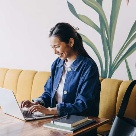 Woman in a denim jacket smiling while working on her laptop, planning her podcast launch services at a cafe table with notebooks nearby.